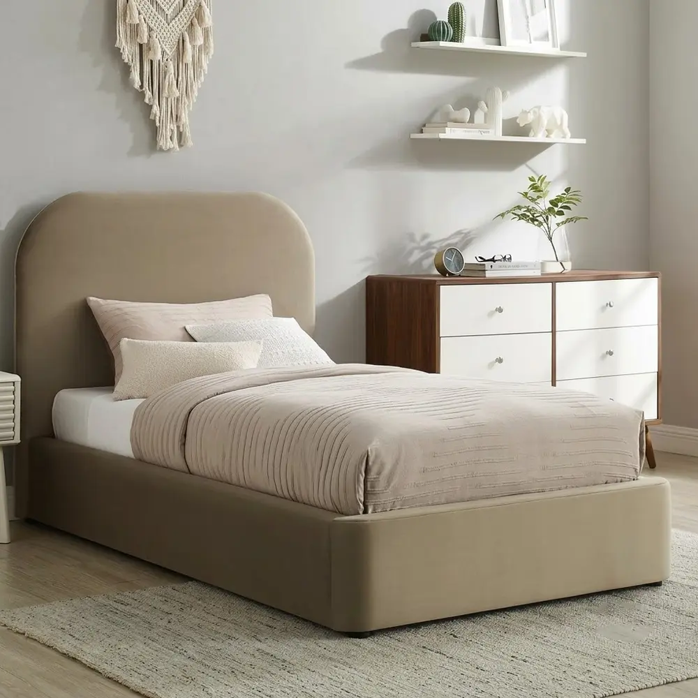 A bed with a tall upholstered headboard and matching footboard in a warm taupe-beige fabric, styled in a modern bedroom with a wood-and-white dresser, floating shelves, and a neutral rug.