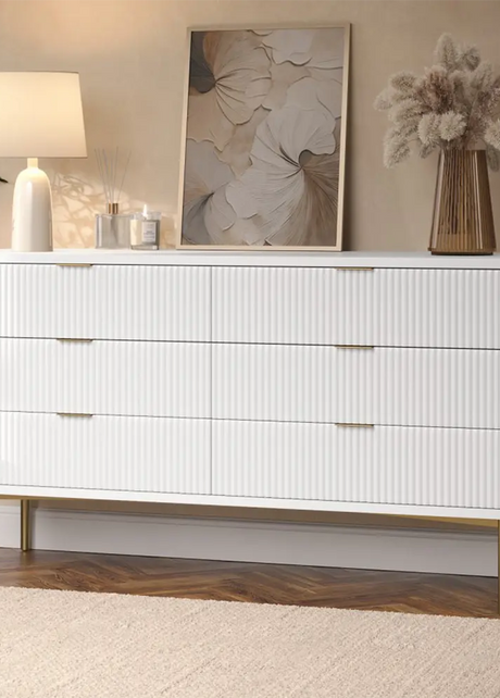 White dresser with gold legs in a room with decor elements like a lamp, painting, and vase.