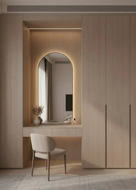A modern built-in vanity area with light wood cabinetry, an illuminated arched mirror, a white desk, a beige upholstered chair, and neutral curtains in a minimalist interior.