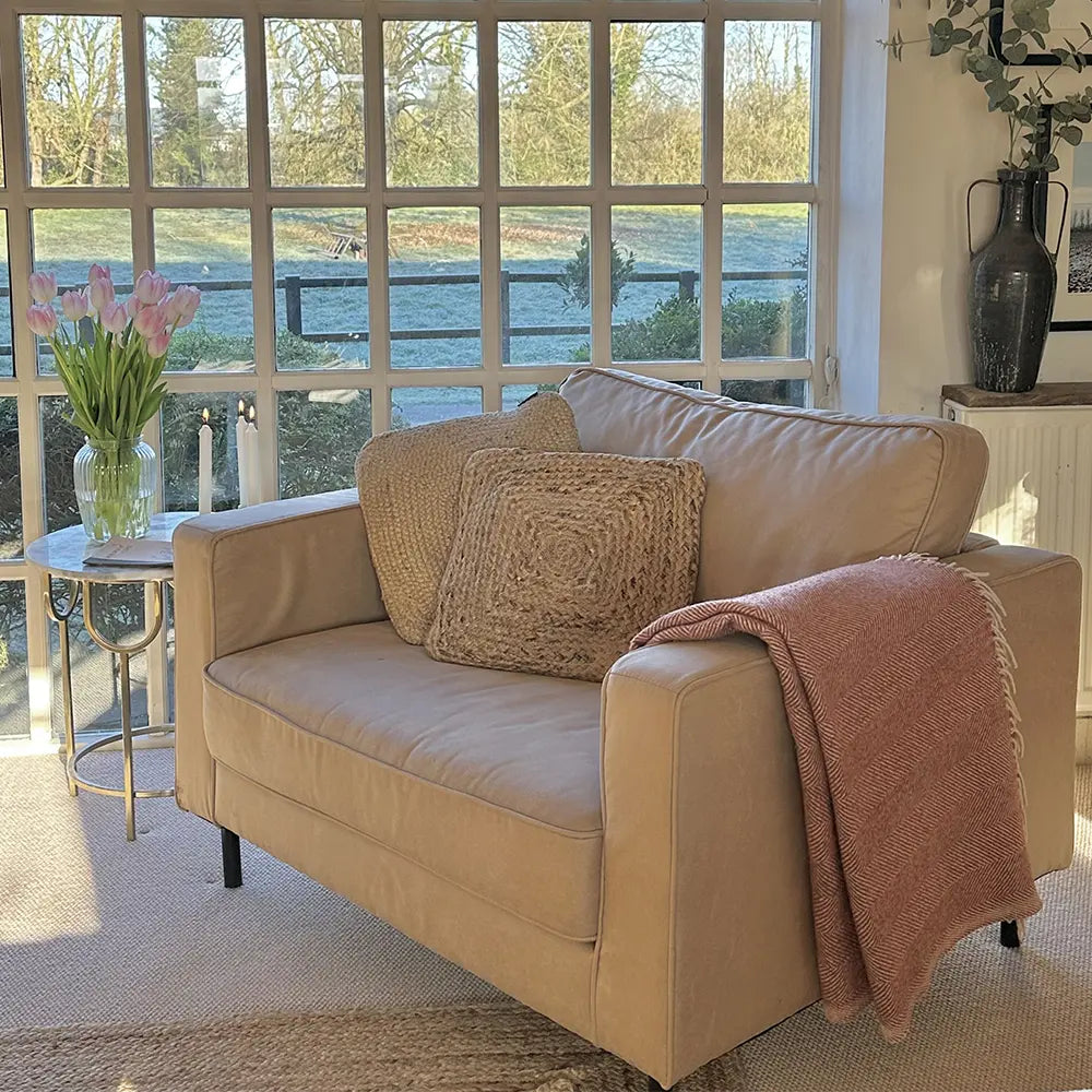 Beige sofa with a pink blanket and woven pillow in a room with large windows.